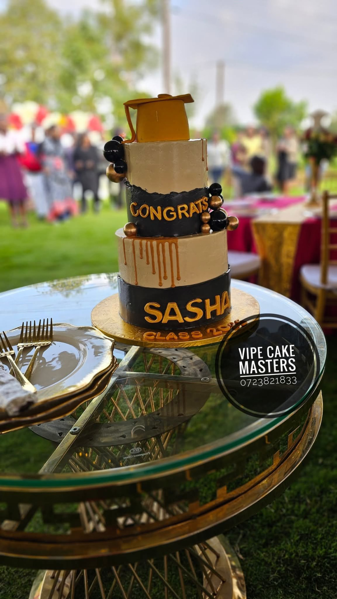 Three-tier cream and black graduation cake with gold drip and Sasha lettering on a glass table at a garden ceremony
