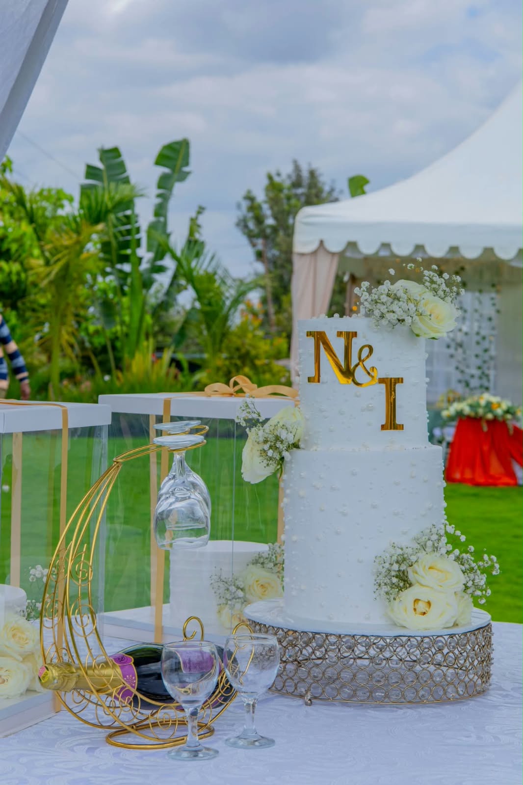 White two-tier pearl wedding cake with gold N & I initials and white roses on a crystal stand in a garden
