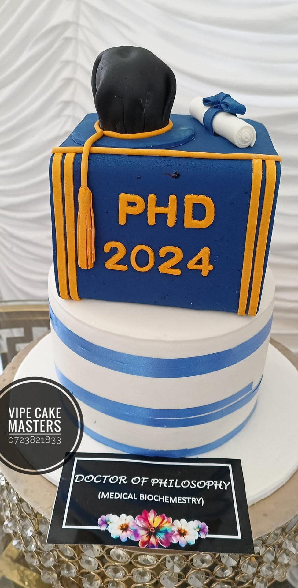 Blue and white Doctor of Philosophy graduation cake with gold lettering, academic cap, tassel and diploma