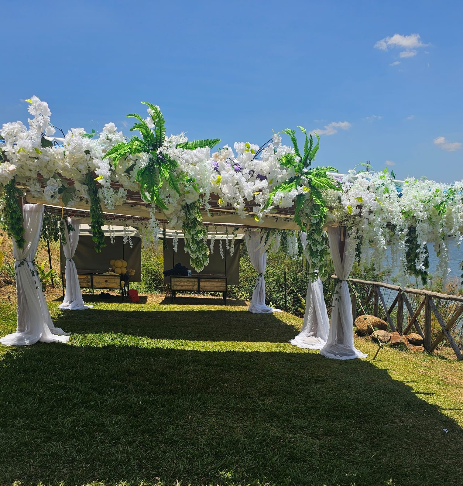 Garden pergola decorated with white flowers, green foliage and white fabric around outdoor lounge seating