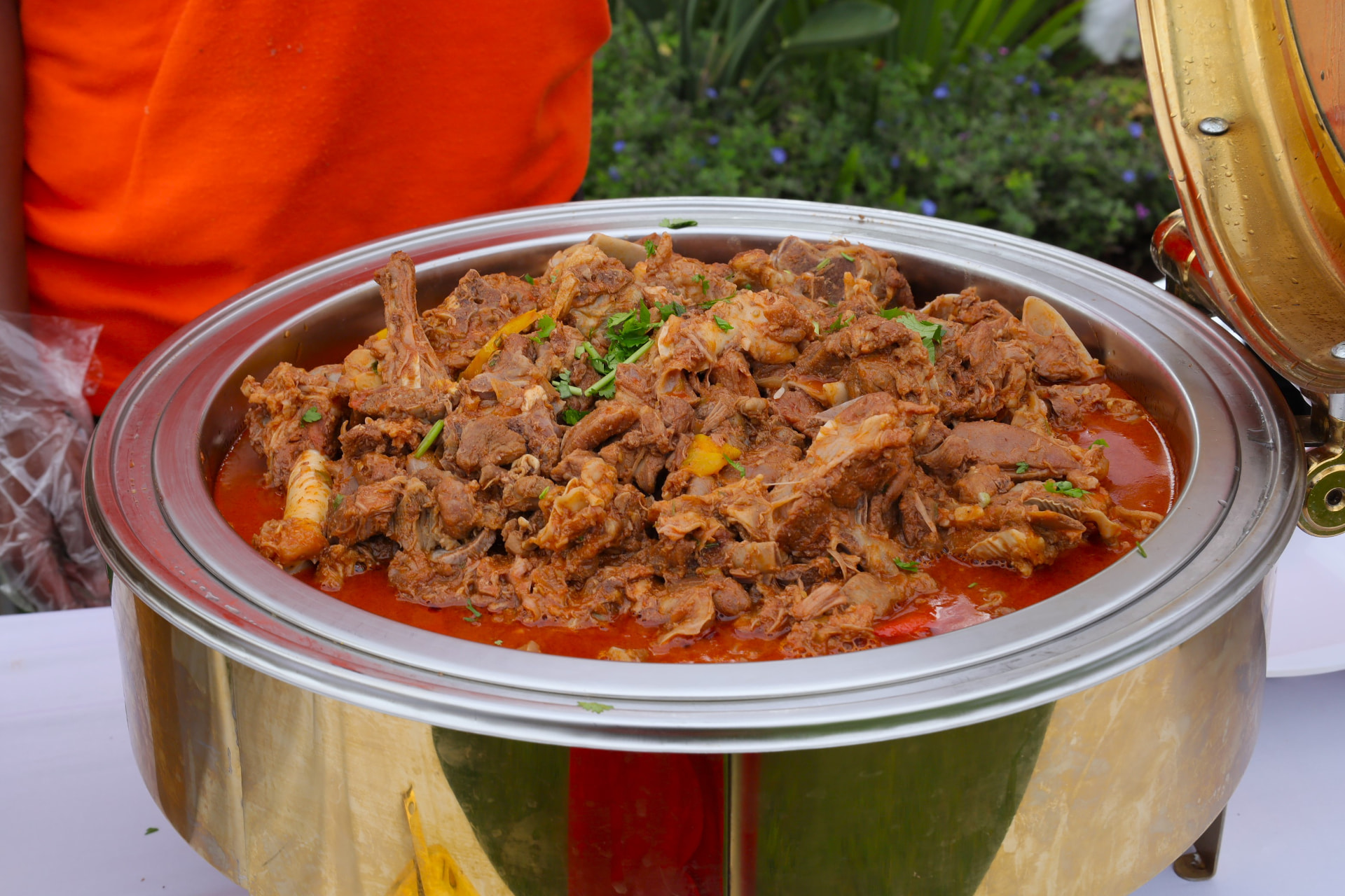 Gold chafer of rich goat meat stew garnished with fresh coriander at an outdoor buffet