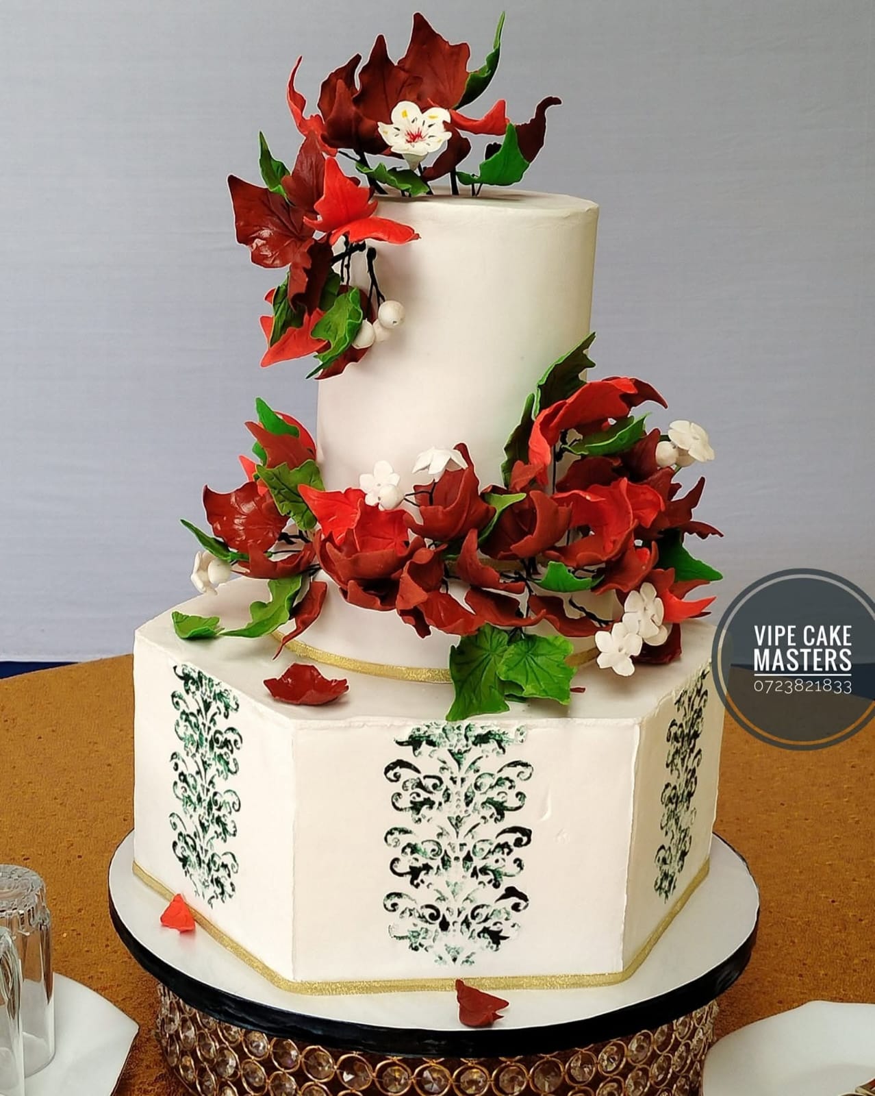 Tall ivory wedding cake with sculpted red autumn leaves, green foliage and white blossoms on a crystal stand