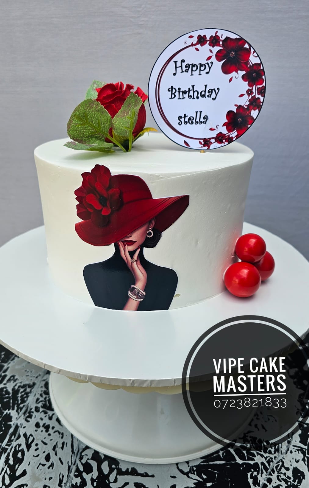 White birthday cake with a lady in a red hat print, red rose, red spheres and Happy Birthday Stella topper