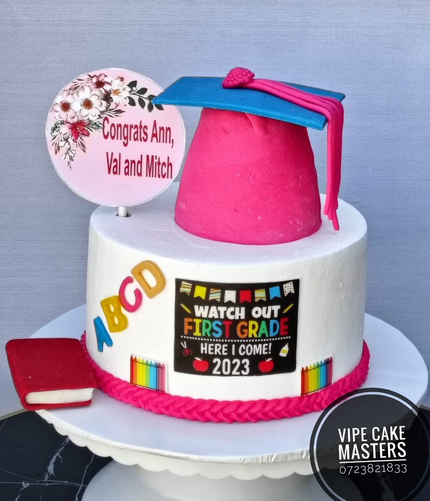 White and pink school celebration cake with graduation cap, alphabet details, book and first grade message