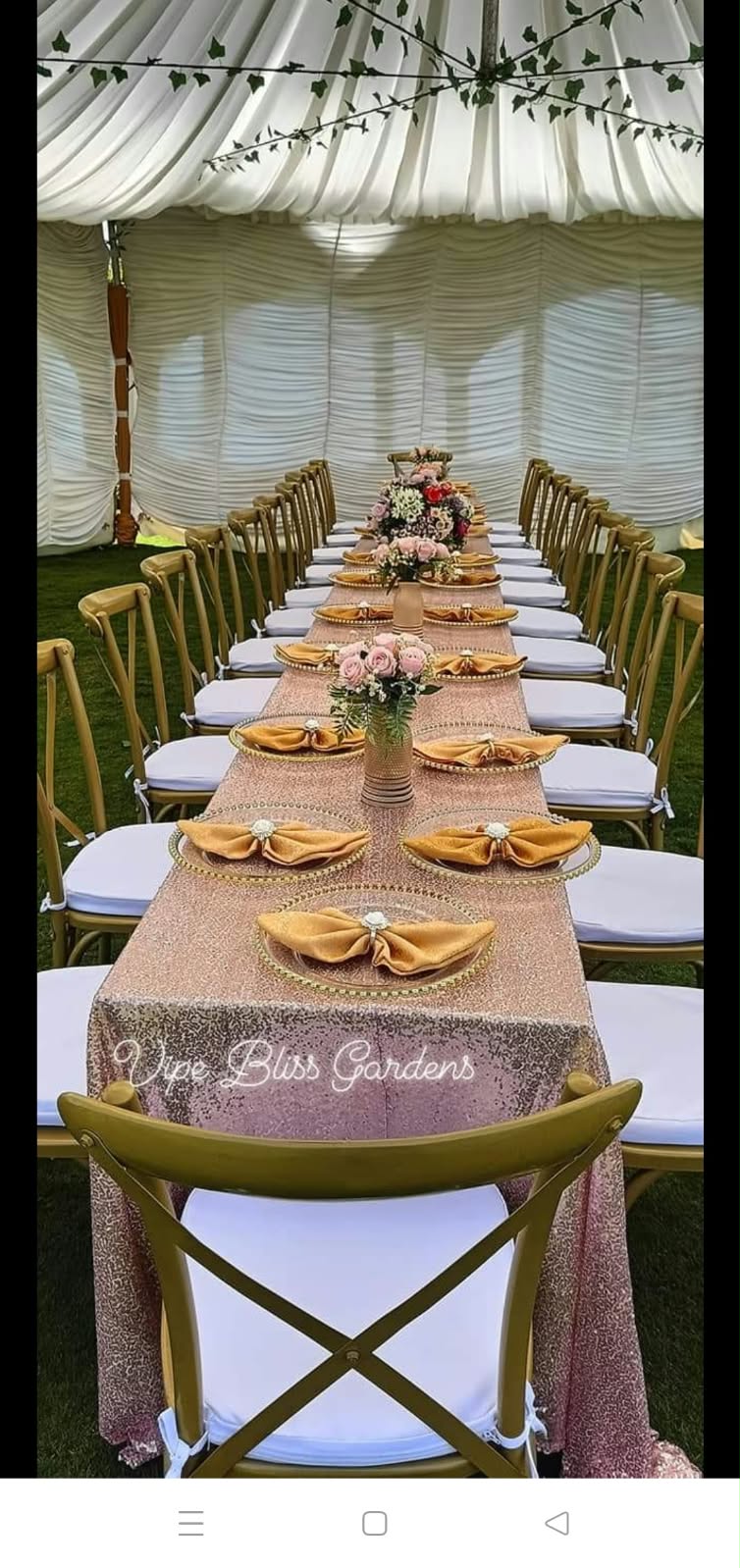Long table with pink sequinned linen, gold place settings, floral centrepieces and gold cross-back chairs beneath white draping