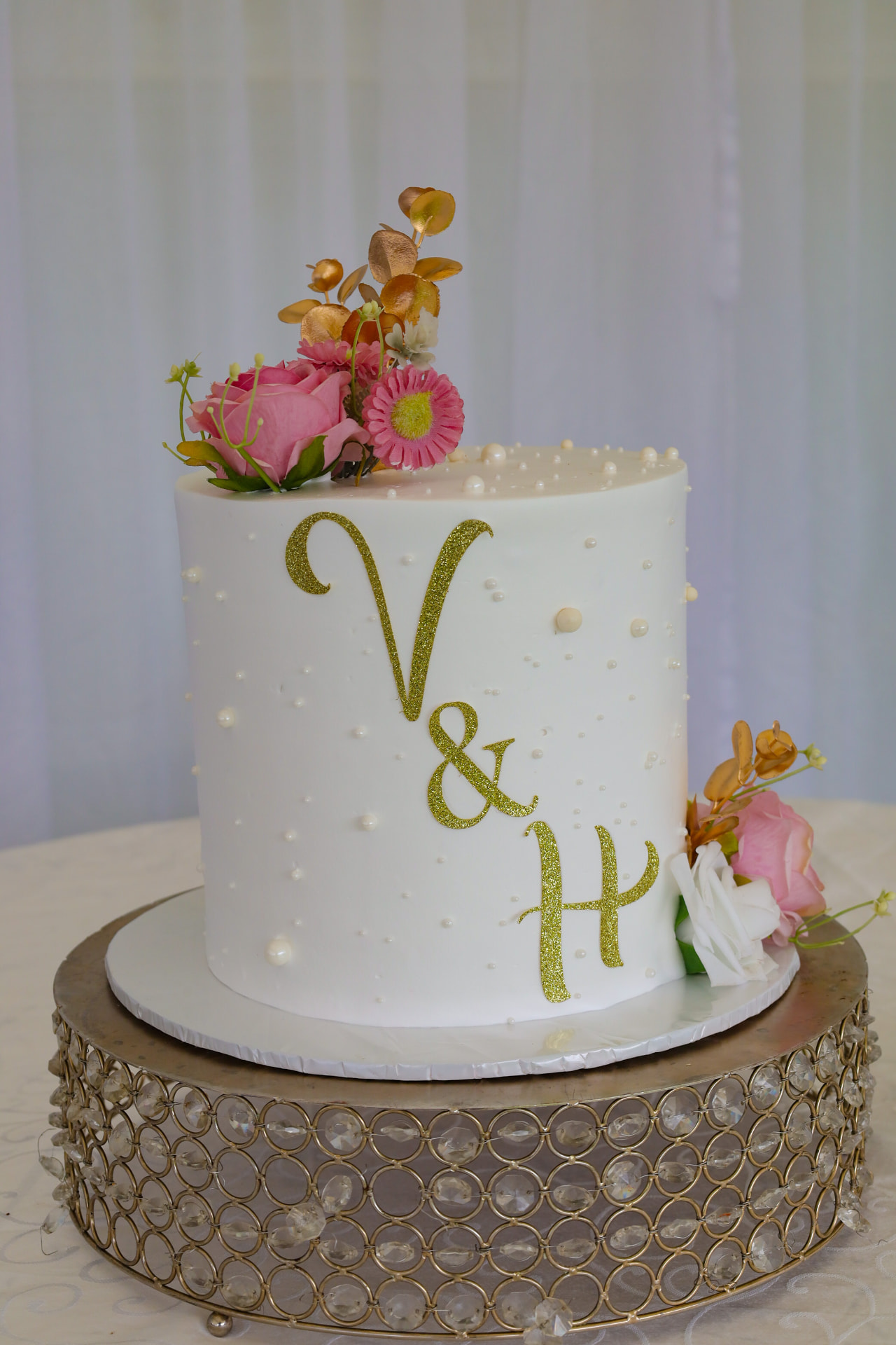 White wedding cake with gold glitter initials, pearls, pink roses and a crystal gold stand