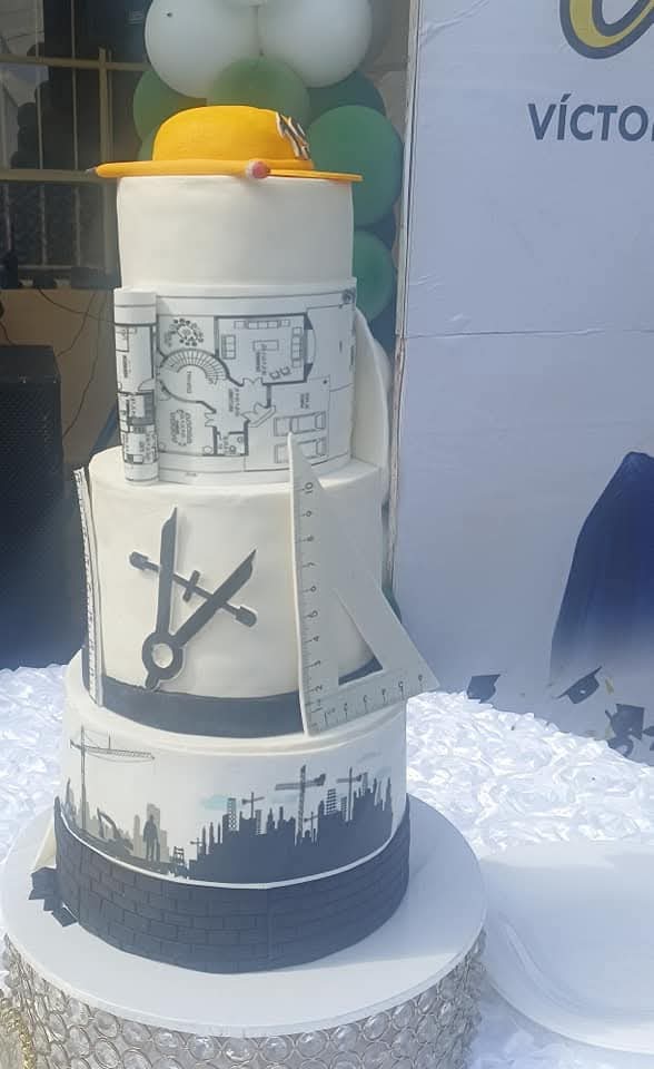 Tall white and charcoal graduation cake decorated with plans, measuring tools, city skyline and a yellow hard hat