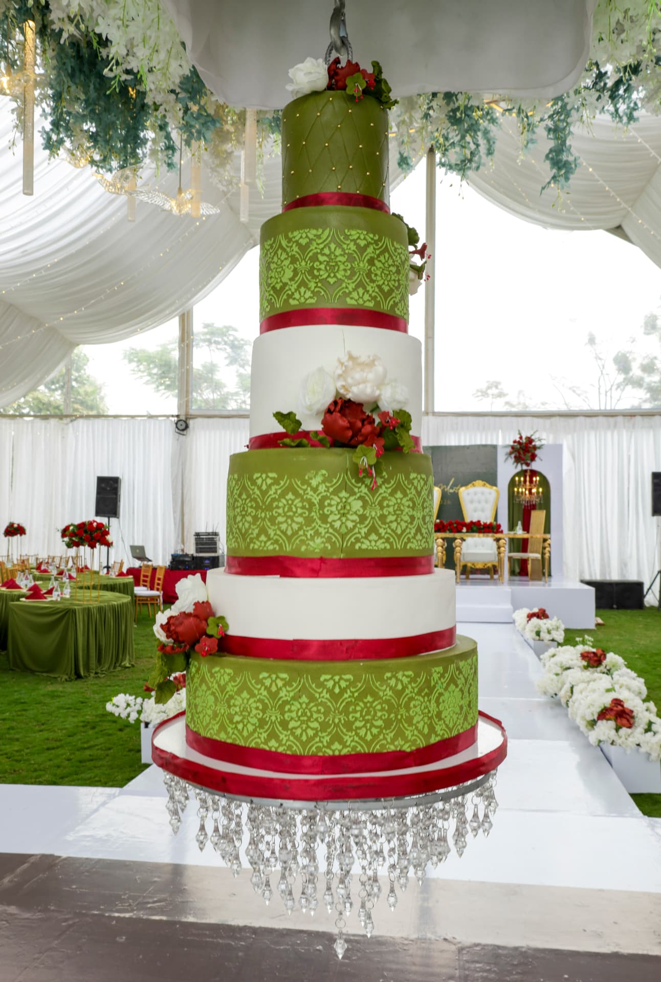 Suspended five-tier green and white wedding cake with red ribbons, lace piping and crystal chandelier drops above a reception aisle