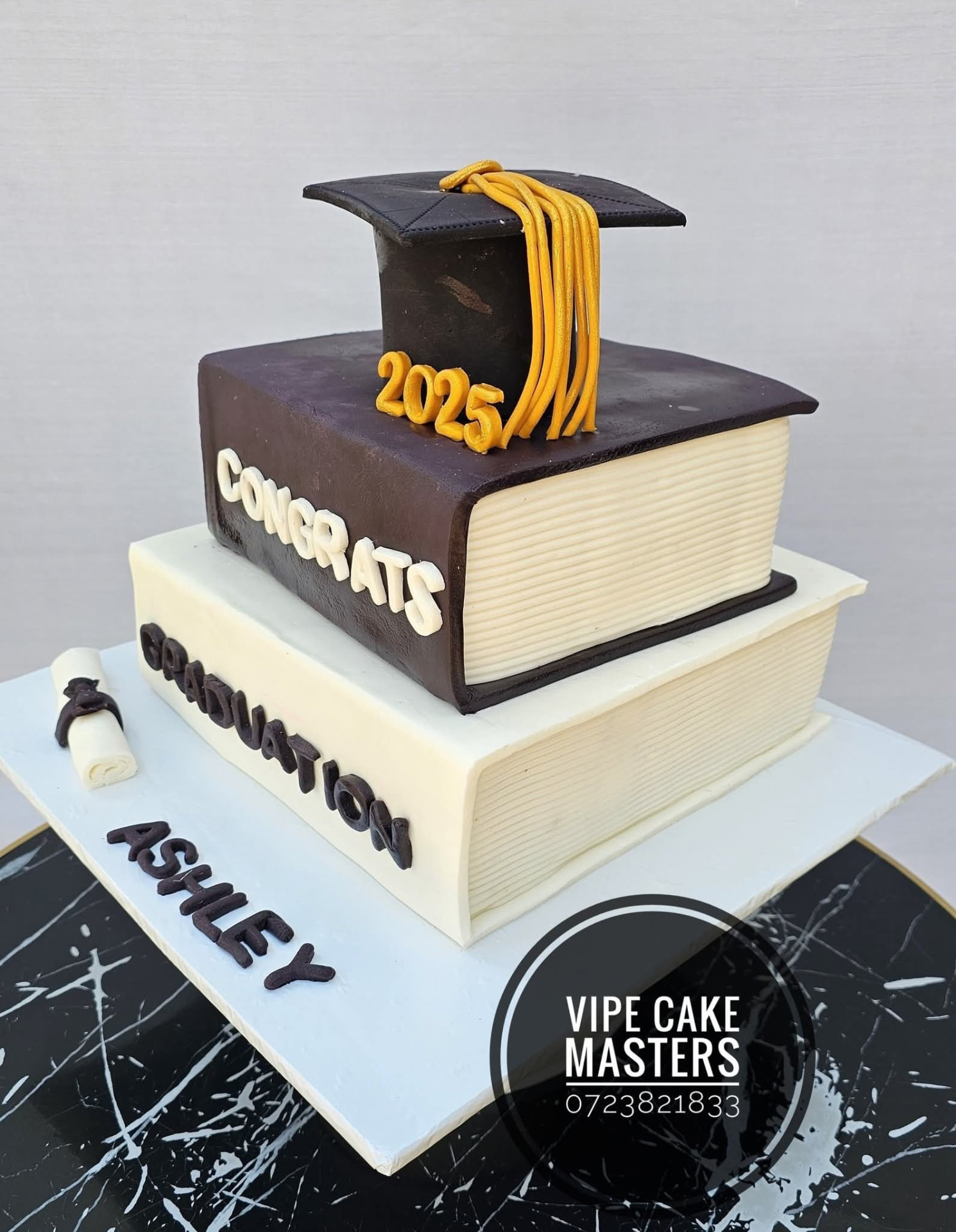 Two-tier graduation cake styled as stacked cream and dark brown books with a black cap and gold 2025 tassel