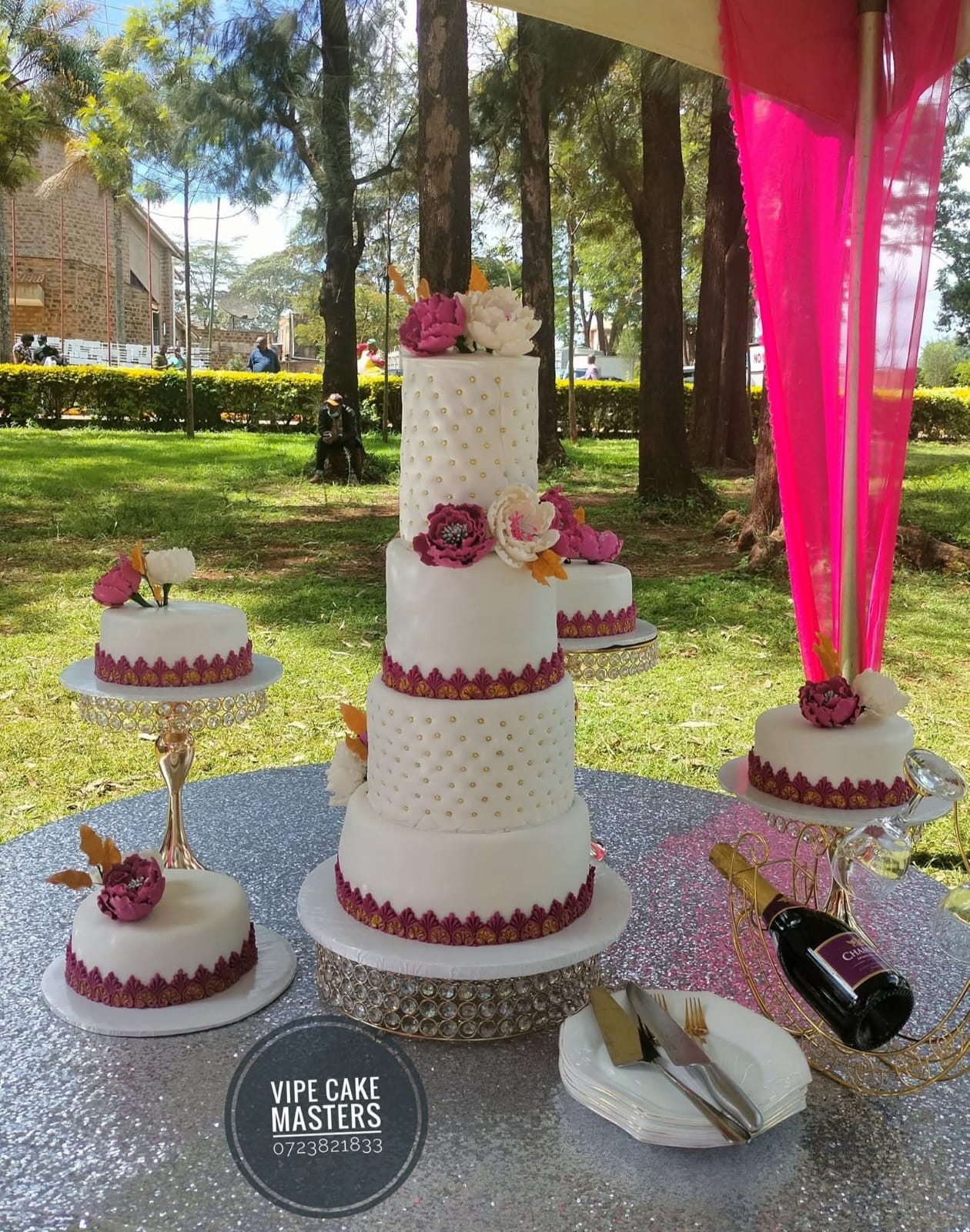 White four-tier wedding cake and matching side cakes with magenta floral borders at an outdoor garden celebration