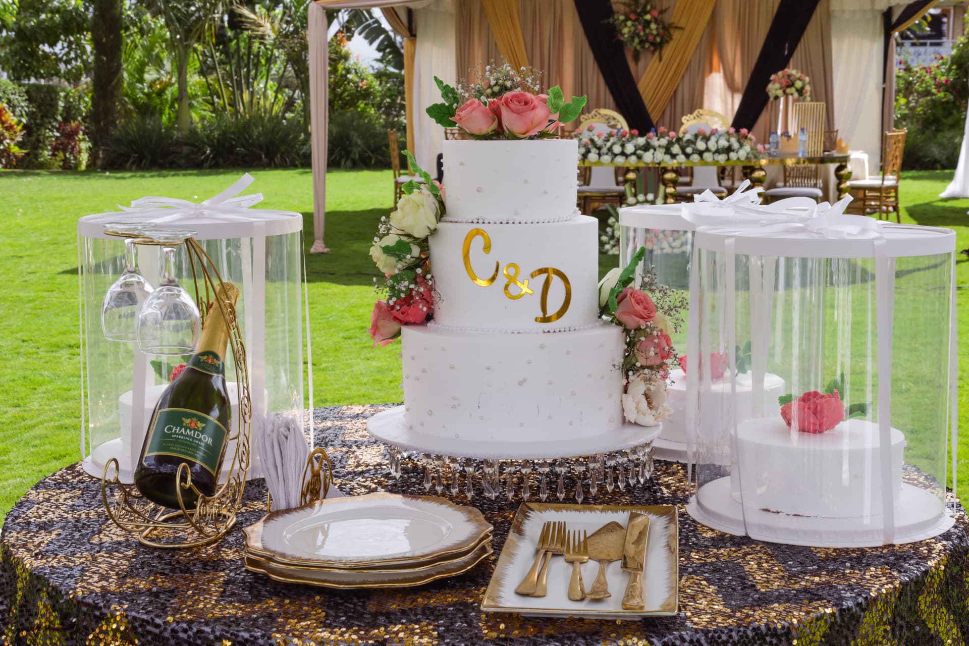 White three-tier Vipe wedding cake with pink roses and gold initials at an outdoor celebration