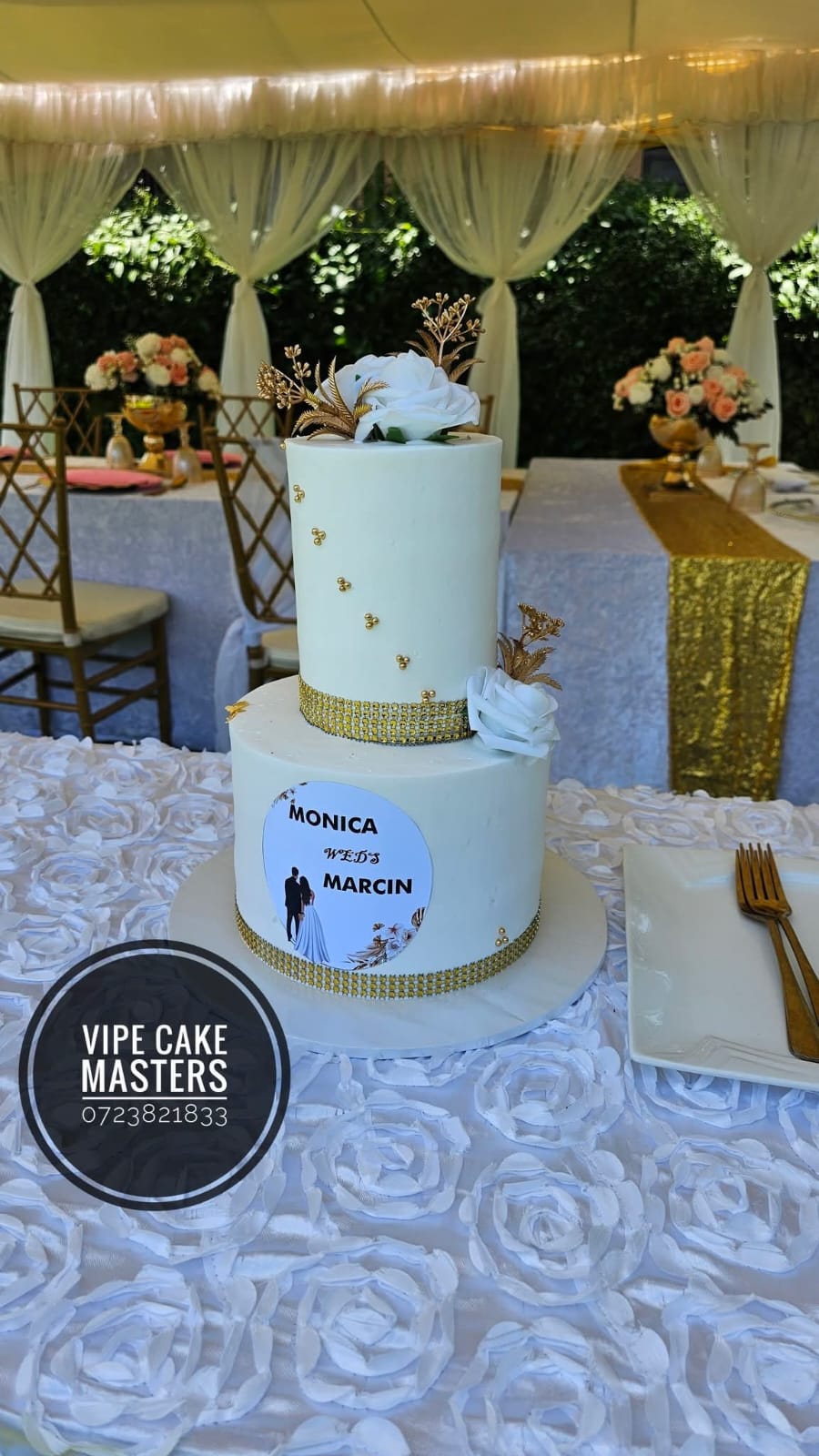 Two-tier white wedding cake with gold crystal bands, white roses and a personalised couple plaque