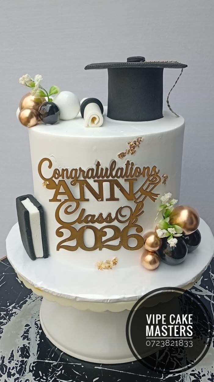 White graduation cake with gold script topper, black cap, sugar book and rose gold and black spheres