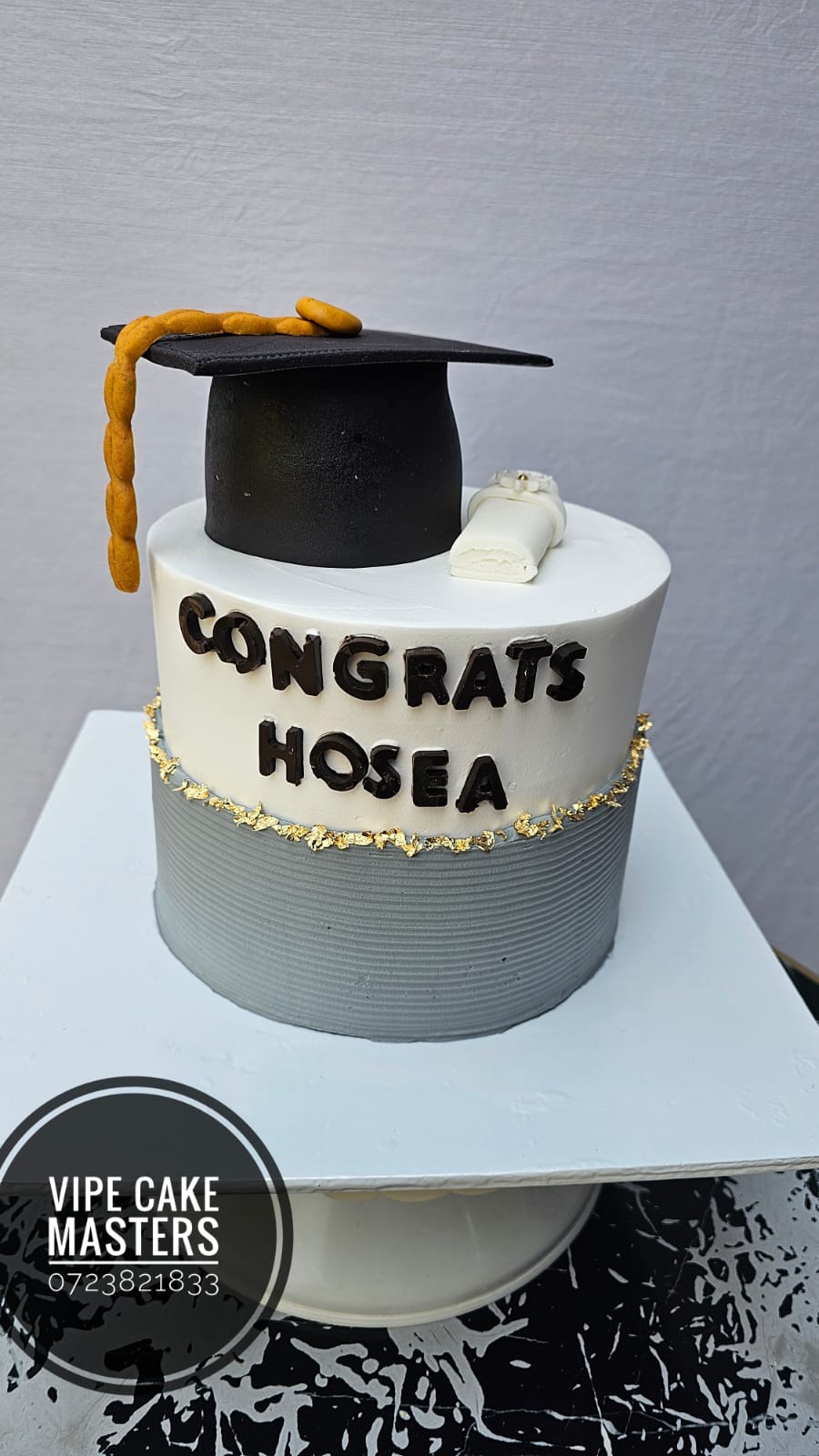 White and grey graduation cake with gold leaf band, black cap, scroll and Congrats Hosea lettering