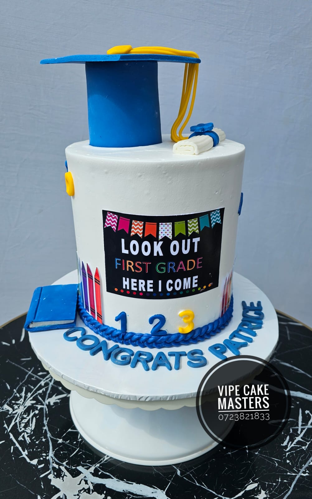 White first grade celebration cake with blue graduation cap, crayons and Congrats Patrice lettering