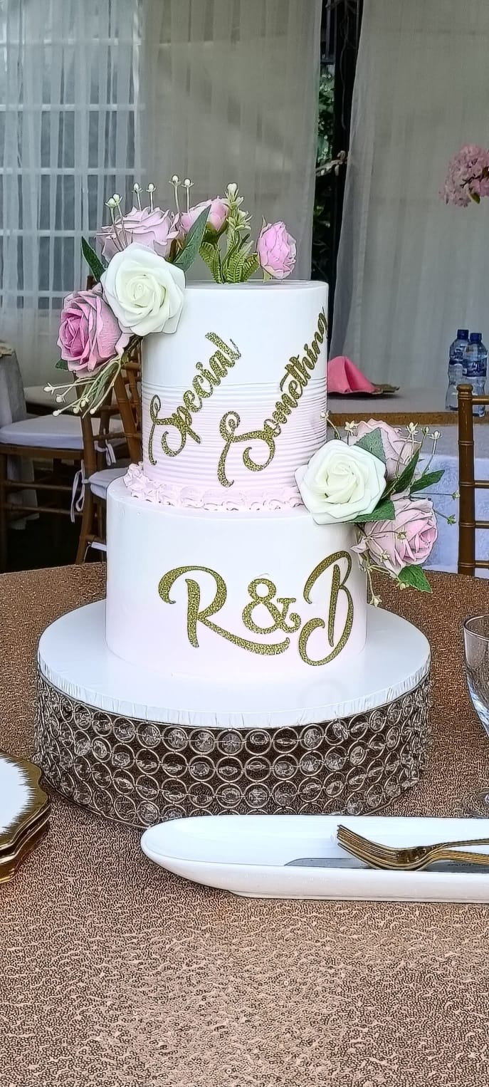 Two-tier white wedding cake with pink and white flowers, gold monogram and personalised couple names on a crystal stand