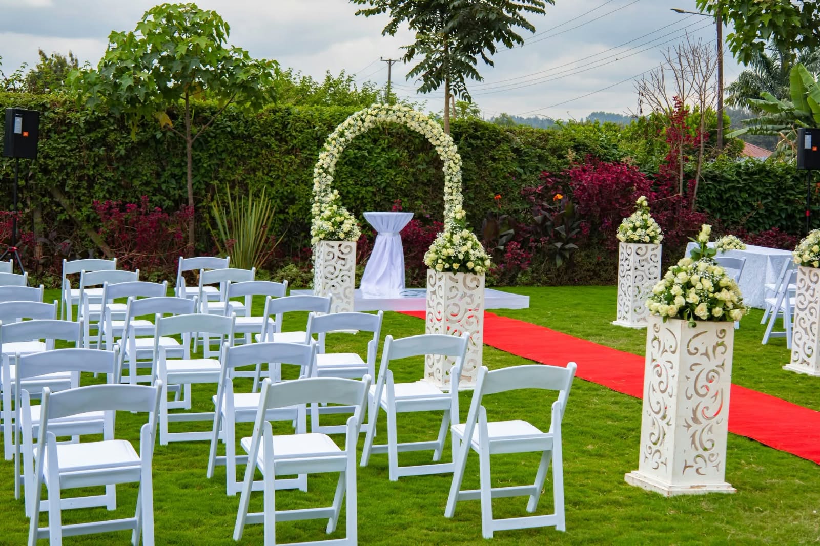 Outdoor garden ceremony with white rose arch, white chairs, carved floral pillars and a red carpet aisle