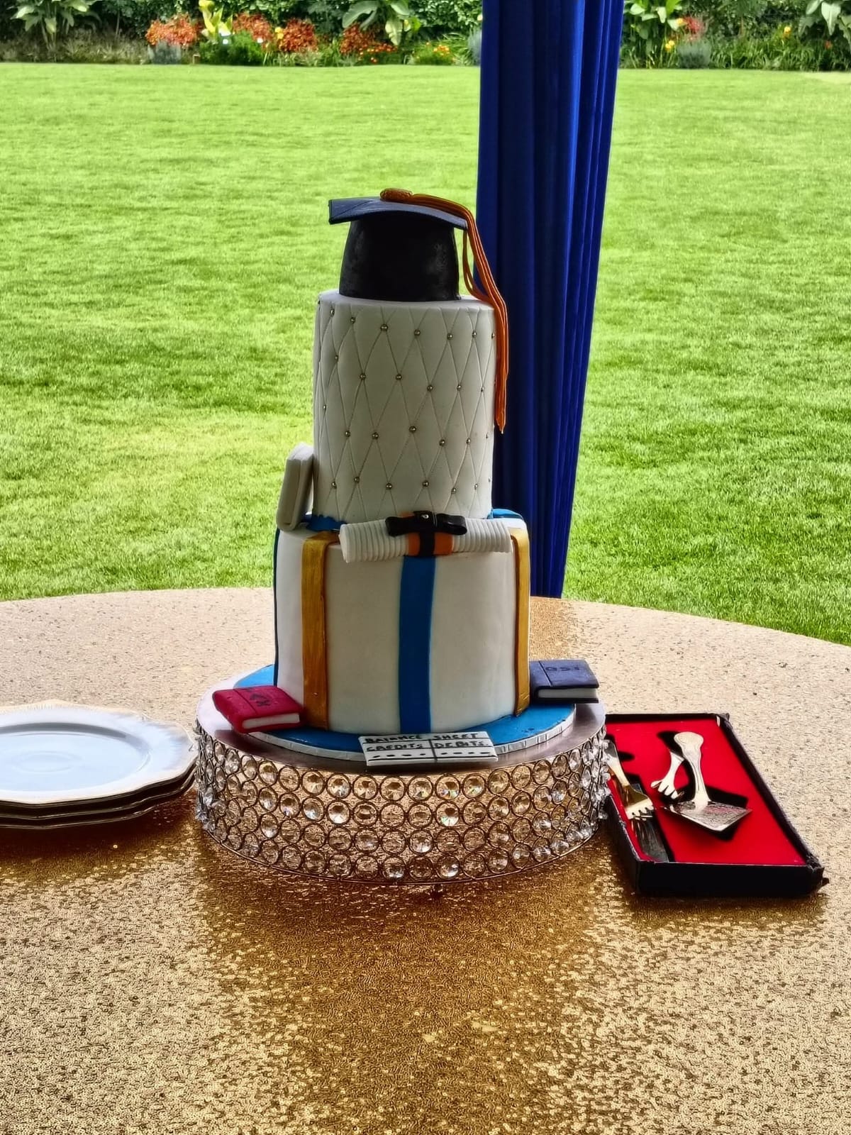 Three-tier graduation cake styled as stacked books with a quilted top tier, black cap, scrolls and writing tools
