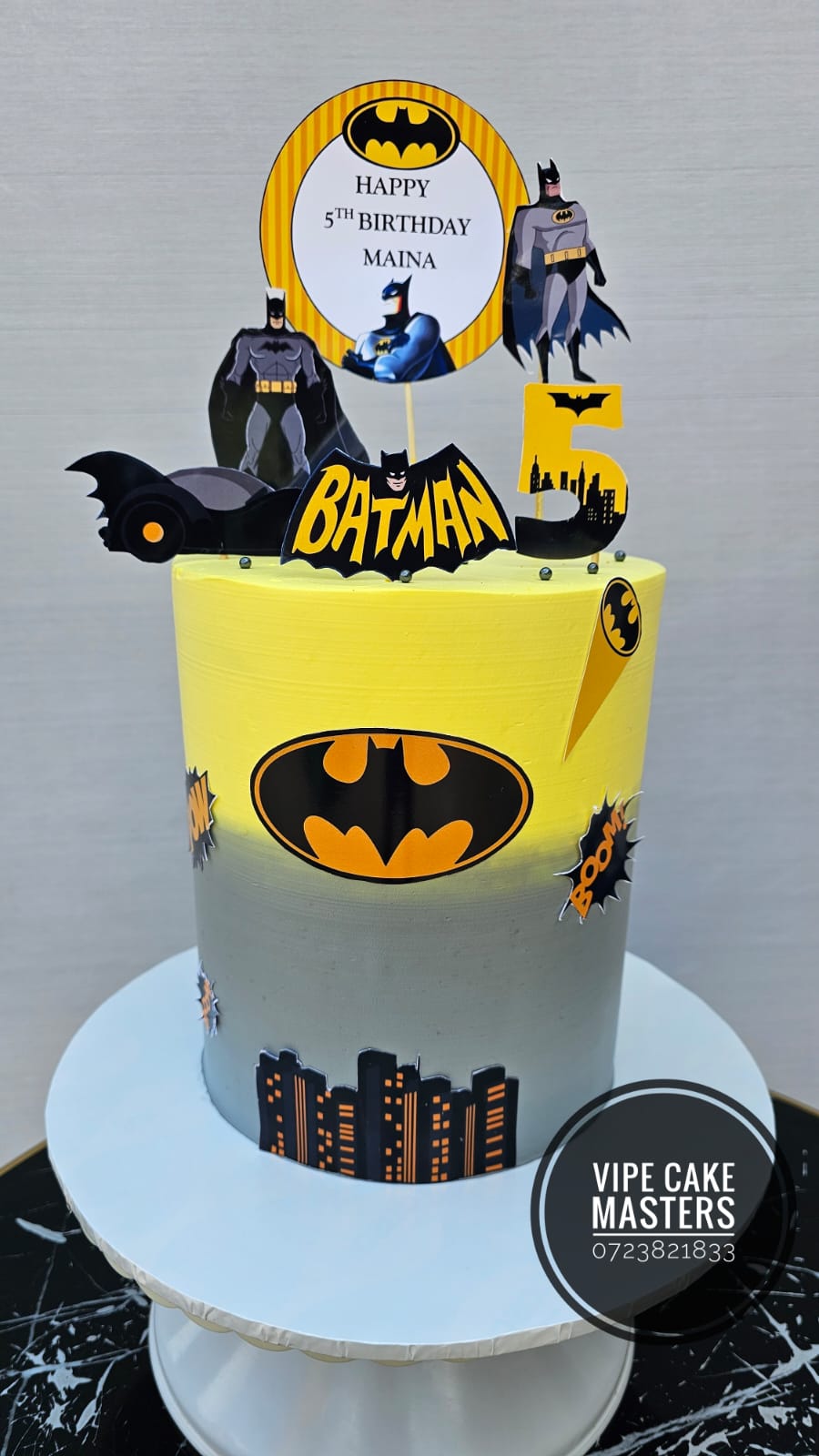 Yellow and grey Batman themed fifth birthday cake with city skyline, bat logo and Happy 5th Birthday Maina topper