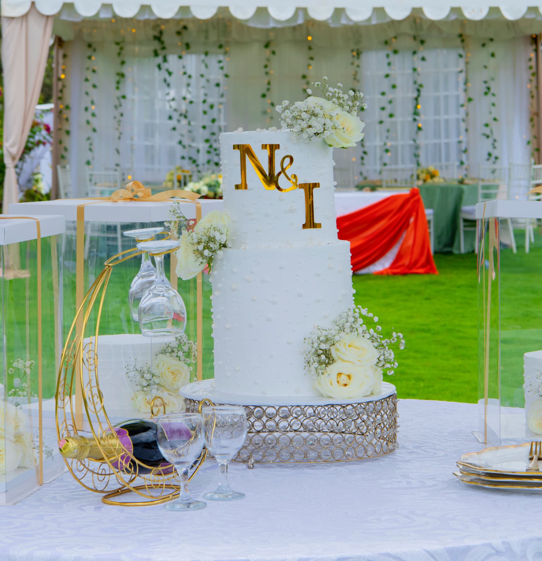 White two-tier wedding cake with gold monogram and white roses at an outdoor garden reception