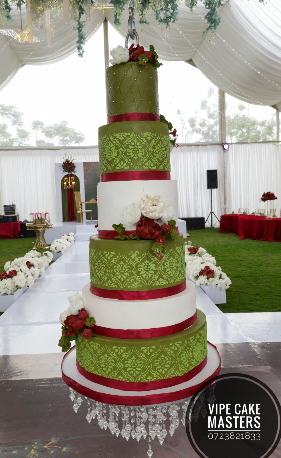 Six-tier green and ivory wedding cake with burgundy ribbons, red and white flowers and a hanging crystal stand