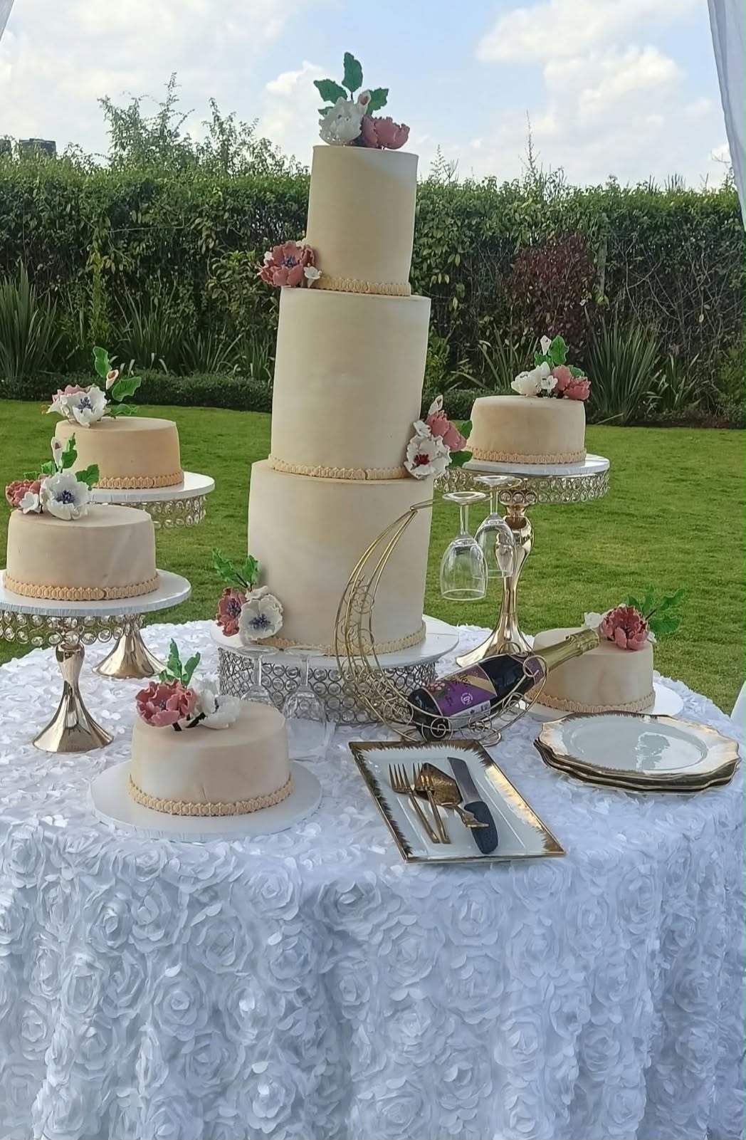 Three-tier ivory wedding cake and five matching side cakes with pink and white flowers on crystal gold stands at a garden reception