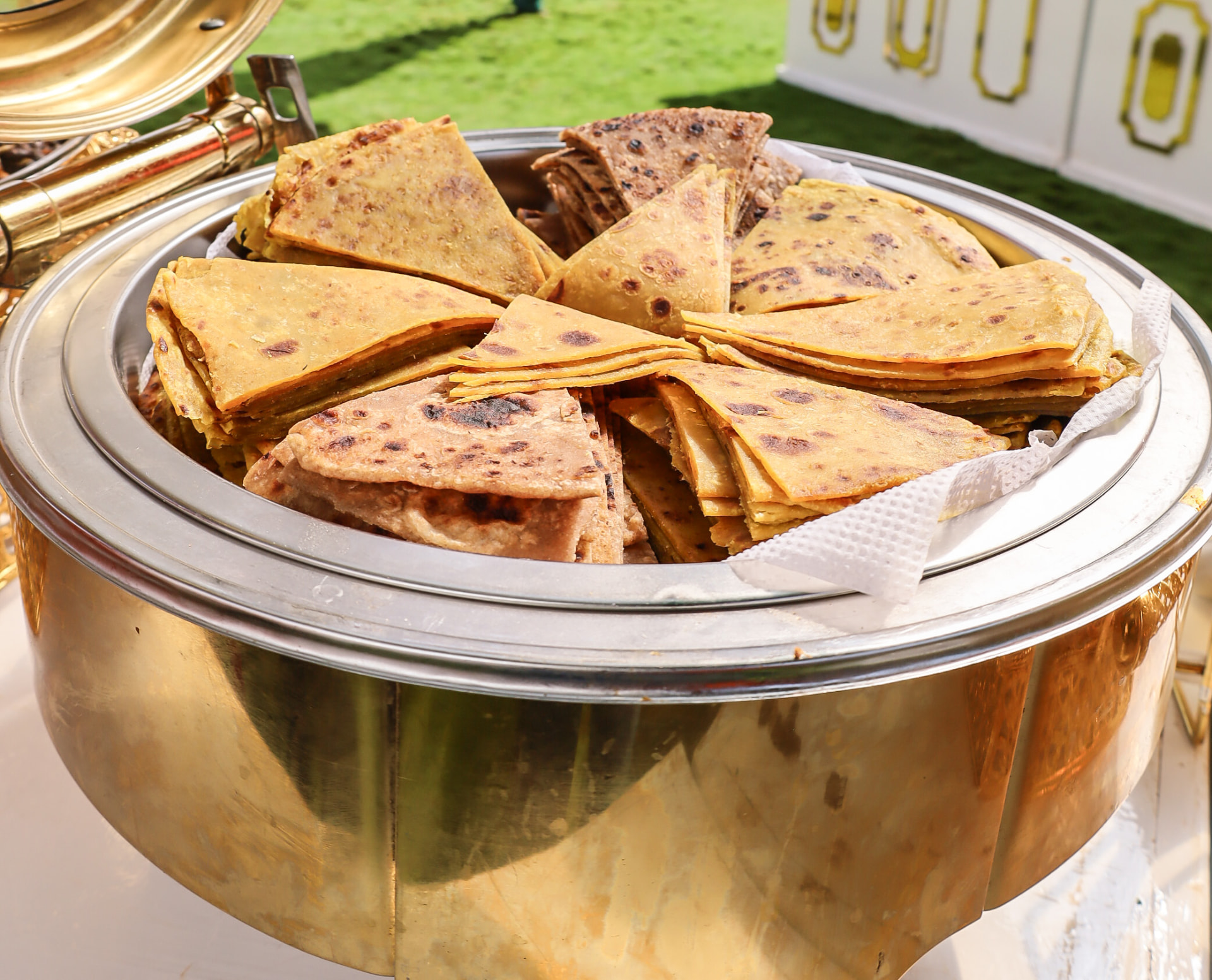 Full gold chafer of chapatis at a garden buffet with white panels and green draping