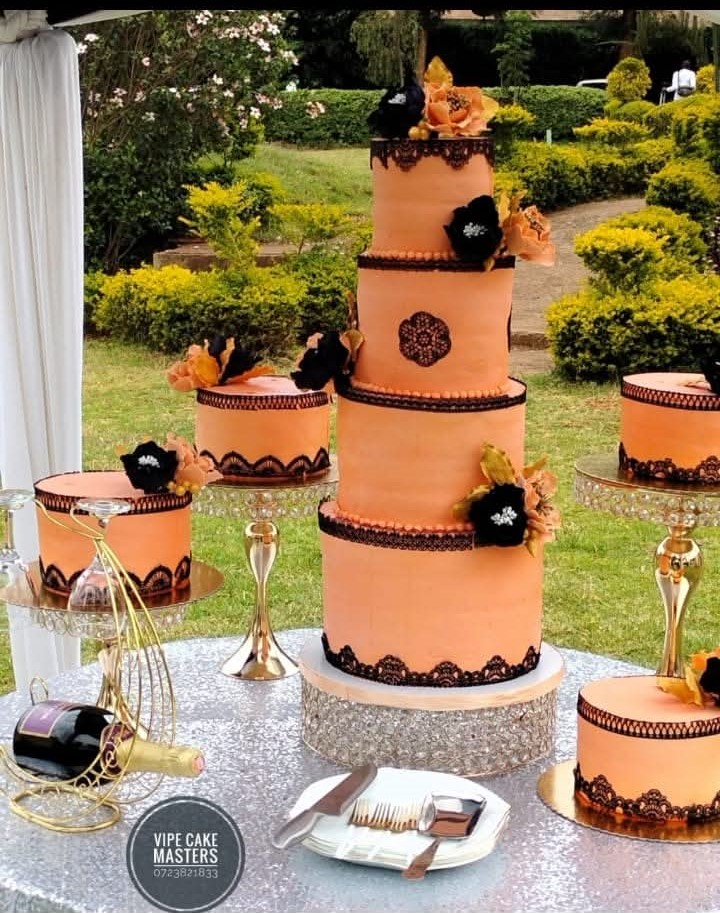 Five-tier peach Vipe wedding cake with black lace and sugar flowers, surrounded by matching side cakes in a garden