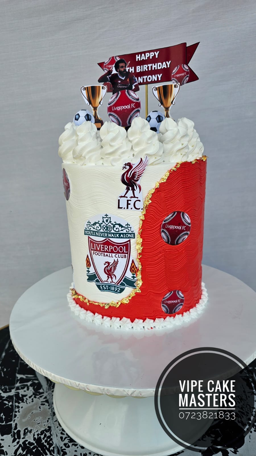 Red and white Liverpool FC themed birthday cake with club crest, trophies, footballs and Happy Birthday Antony banner